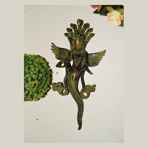 Może przedstawiać: Metalowa statua w kolorze zielonym i złotym przedstawiająca bóstwo z wieloma głowami węża i skrzydłami. Bóstwo trzyma w dłoniach mały przedmiot.