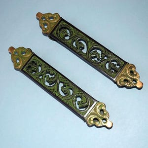 May include: Two decorative cabinet pulls with a green and black scroll design. The ends are gold-colored with ornate detailing. The pulls are rectangular and are approximately 6 inches long.