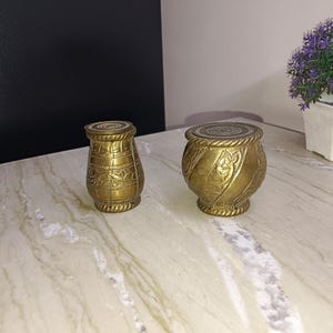 Puede incluir: Dos recipientes decorativos antiguos de latón. El recipiente de la izquierda tiene forma de jarrón con diseños florales grabados. El recipiente de la derecha tiene forma redonda con diseños florales grabados. Ambos recipientes son de color dorado.