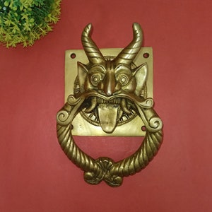 May include: A brass door knocker depicting a devil's face with horns, a protruding tongue, and an open mouth. The knocker has a ring-shaped handle and is mounted on a square plate. The design is detailed and ornate, with a vintage aesthetic.