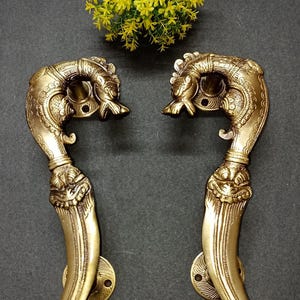May include: A pair of gold-coloured metal door handles featuring intricate embossed carvings of mythical creatures. Each handle has a curved top section shaped like a head and a long, smooth lower grip area with circular mounting plates at both ends.
