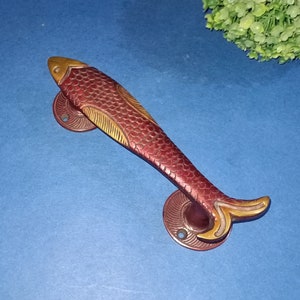 May include: A red and gold fish-shaped door handle with a detailed design. The handle is made of metal and has two mounting plates.