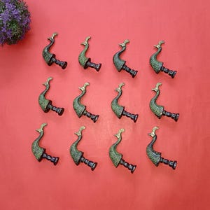 Peut inclure: Douze bouchons de bouteille décoratifs en forme de paon. Chaque bouchon présente un corps vert avec des accents dorés et une base noire. Ils sont disposés sur une surface rouge, avec un petit arrangement floral violet en haut à gauche.