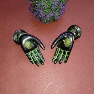 May include: Two black metal hands with green accents and gold details. The hands are open and facing upwards, as if offering something.