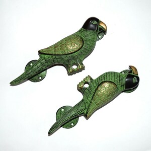 Może przedstawiać: Para zielonych i złotych uchwytów do drzwi w kształcie papugi. Uchwyty są wykonane z metalu i mają szczegółowy wzór.