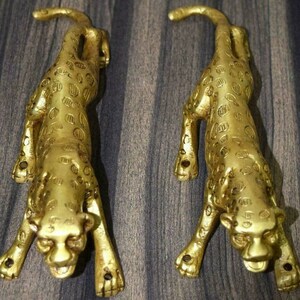 May include: Two gold-colored metal leopard figurines. The leopards are in a crouching position with their heads down and their tails curled up. The leopards are detailed with spots and have a realistic appearance.