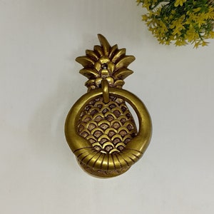 May include: A gold-colored pineapple door knocker. The knocker features a detailed pineapple design with a ring handle. The pineapple has a textured surface and is made of metal. The door knocker is a decorative item.