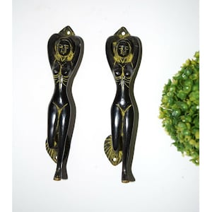 Può includere: Un paio di maniglie per porte in metallo nero a forma di corpo di donna. Le maniglie hanno una finitura dorata e sono in posizione eretta.