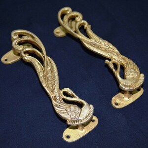 May include: Two gold-toned metal door handles shaped like peacocks with intricate details. The handles are designed with a long, curved shape and a decorative base.