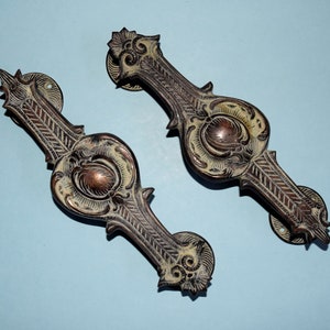 May include: Two ornate brass door handles with intricate floral designs. The handles are a dark brown color with a light brown patina.