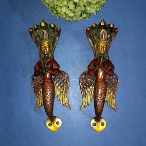May include: A pair of ornate brass door handles shaped like a mermaid with wings. The mermaids are holding their hands together in a prayer position. The handles are painted in shades of gold, red, and green.
