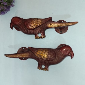 Może przedstawiać: Dwa dekoracyjne uchwyty do drzwi w kształcie papugi. Uchwyty wykonane są z czerwonobrązowego metalu ze złotymi akcentami na skrzydłach. Każdy uchwyt ma okrągłą podstawę do montażu i otwór na śrubę.