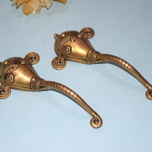 May include: Two gold-toned metal door handles shaped like elephant heads with intricate details. The handles have a curved design and are perfect for adding a unique touch to any home.