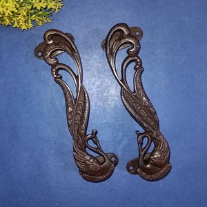 Może przedstawiać: Para ozdobnych, ciemnobrązowych metalowych klamki do drzwi w kształcie pawia z misternymi detalami. Klamki są zaprojektowane z głową i szyją ptaka, a ciało ptaka tworzy sama klamka.