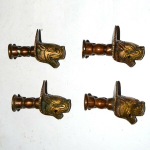 Könnte beinhalten: Vier antike Gardinenstangen-Endstücke aus Bronze. Jedes Endstück zeigt einen detaillierten, stilisierten Tierkopf mit einem markanten Ohr und einem zylindrischen Stangenanschluss. Die Köpfe sind goldfarben mit grünen Akzenten, die Stangen sind dunkler Bronze.