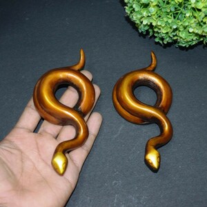 Könnte beinhalten: Ein Paar goldfarbene, schlangenförmige Ohrdehner. Die Dehnungen sind aus einem glatten, glänzenden Material gefertigt und haben ein realistisches Schlangendesign.