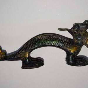 May include: A black and green dragon door handle with gold accents. The handle is shaped like a dragon with its head turned to the side and its tail curled up. The handle is made of metal and has a detailed design.