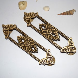 May include: Two ornate brass door pulls with a floral design. The pulls are rectangular with a decorative butterfly design at each end.