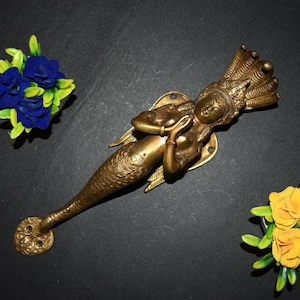 Puede incluir: Una manilla de puerta de latón dorado con forma de sirena con una corona y alas de serpiente. La sirena tiene las manos juntas en posición de oración.