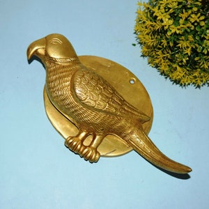 Könnte beinhalten: Ein goldfarbener Türklopfer aus Messing in Form eines Papageis mit einem detaillierten Design. Der Papagei sitzt auf einer runden Basis.