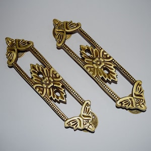 Puede incluir: Dos tiradores de puerta de metal dorado adornados con un diseño floral. Los tiradores tienen una forma rectangular con un marco decorativo y un extremo en forma de mariposa.