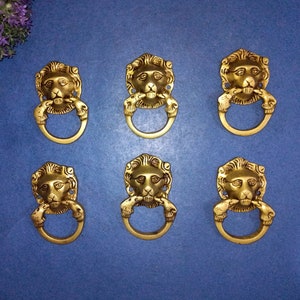 Pode incluir: Seis puxadores de gaveta de cabeça de leão dourados com uma alça de anel. As cabeças de leão são detalhadas com uma juba e uma boca fechada.