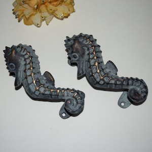 Op de afbeelding: Twee antieke bronzen deurgrepen in de vorm van zeepaardjes met een verweerde afwerking. De grepen zijn gedetailleerd met getextureerde schubben en hebben een ring aan de onderkant voor montage.