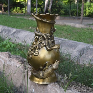 May include: A gold metal vase with intricate carvings of snakes and flowers. The vase has a wide mouth and a round body.