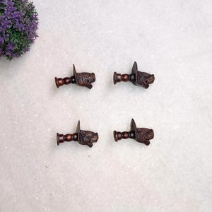 Op de afbeelding: Vier kleine, donkerbruine decoratieve ladeknopjes. Elk knopje heeft een gesneden dierenkopontwerp. Ze zijn gerangschikt in een vierkant patroon op een licht oppervlak, met een paarse bloemstuk in de linkerbovenhoek. Elk knopje is ongeveer 5 cm lang.