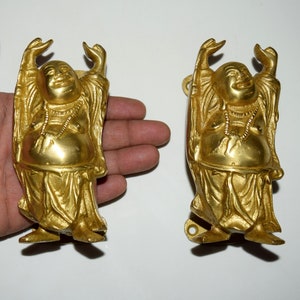 以下が含まれることがあります： 腕を上げた笑顔の仏像が2体。金色で、白い背景に立っています。
