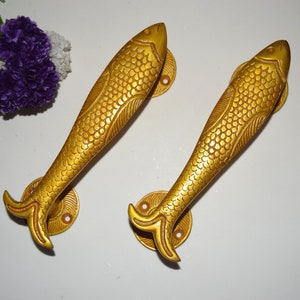 May include: Two gold-toned fish-shaped door handles with detailed scales and a textured finish. The handles are mounted on round bases with decorative edges.