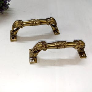 Könnte beinhalten: Zwei antike Türgriffe aus Messing. Jeder Griff hat an jedem Ende ein Elefantenkopf-Design und verzierte Details entlang der Stange. Die Griffe sind auf rechteckigen Platten mit Schraubenlöchern montiert, vor weißem Hintergrund.