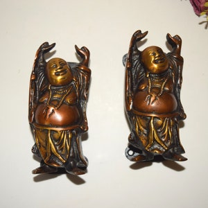 May include: Two gold and bronze coloured door handles shaped like a laughing Buddha with raised arms. The handles are mounted on a white background.
