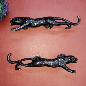 Op de afbeelding: Twee zwarte metalen kastgrepen in de vorm van een luipaard. De grepen hebben een gevlekt patroon en staan in een rennende houding. De grepen zijn gemonteerd op een roodbruin oppervlak.