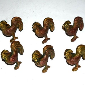 May include: Six bronze-colored rooster figurines with detailed feather and comb designs. The roosters are posed in a standing position, with a mix of gold, green, and red-brown hues. The figurines are arranged on a white background.