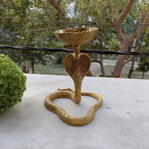 Könnte beinhalten: Ein goldfarbener Metall-Räucherofen in Form einer Kobra mit filigranen Details. Der Ofen hat eine runde Oberseite zum Aufstellen von Räucherstäbchen und einen gewundenen Körper.