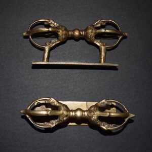 May include: Two gold-coloured metal Tibetan prayer wheels with intricate designs, mounted on a rectangular base.