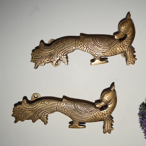 May include: Two antique brass-coloured decorative duck-shaped door pulls. Each pull features intricate engraved details, including feathers and a stylised head. The pulls are mounted on a white surface, with a small purple plant in the background.