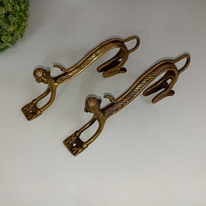 May include: Two ornate brass hooks shaped like stylized animals with curved necks and detailed bodies. The hooks are designed to be mounted on a wall.
