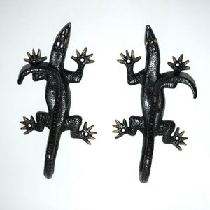 Op de afbeelding: Twee zwarte metalen haken in de vorm van een hagedis met gouden accenten. De hagedissen kijken elkaar aan met hun staarten omhoog gekruld.