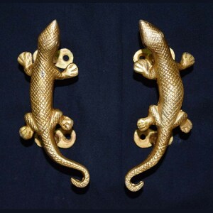 Peut inclure: Une paire de poignées de porte en métal doré en forme de lézards. Les poignées ont une surface texturée et sont conçues pour être montées sur une porte.