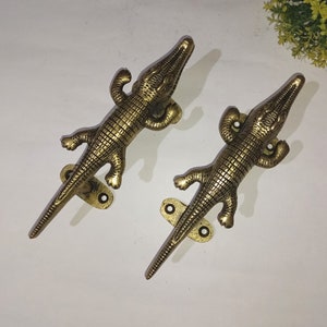 May include: Two gold-toned crocodile door handles with detailed scales and a textured finish. Each handle has a metal plate with two holes for mounting.