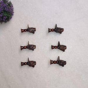 Könnte beinhalten: Sechs bronzefarbene Schubladengriffe mit Löwenkopf-Design. Jeder Griff hat einen dekorativen Stiel und einen kleinen, spitzen Griff. Die Griffe sind auf einer hellgrauen Oberfläche angeordnet, mit einer lila Blumengesteck oben links.