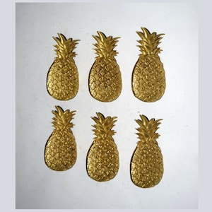 Op de afbeelding: Zes goudkleurige decoratieve voorwerpen in de vorm van een ananas. De ananas zijn allemaal even groot en even gevormd en zijn gerangschikt in twee rijen van drie.
