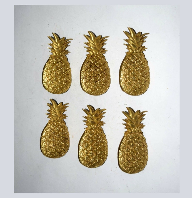 May include: Six gold-colored pineapple-shaped decorative objects. The pineapples are all the same size and shape, and they are arranged in two rows of three.