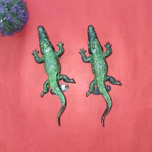 May include: A pair of green alligator-shaped door handles with a textured finish. The handles are made of metal and have a dark green and bronze colour scheme. The alligator door handles are mounted on a red background.