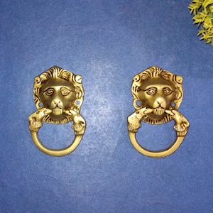 May include: Two gold lion head door knobs with a ring handle. The lion heads are detailed with a textured mane and a realistic expression.