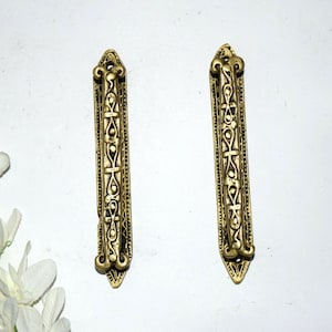 May include: Two ornate gold-toned metal door handles with intricate designs. The handles are rectangular with a pointed top and bottom.