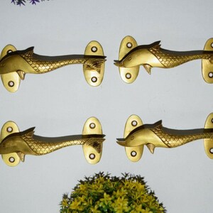 Puede incluir: Cuatro tiradores de puerta dorados en forma de pez con escamas. Cada tirador tiene una base redonda de forma ovalada.