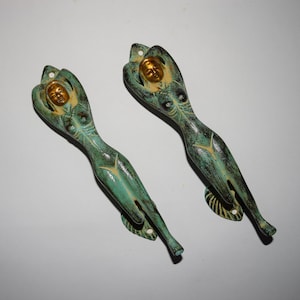 Puede incluir: Dos tiradores de puerta de estilo antiguo con forma de figura femenina reclinada. Los tiradores están hechos de metal y tienen un acabado de pátina verde con detalles dorados.